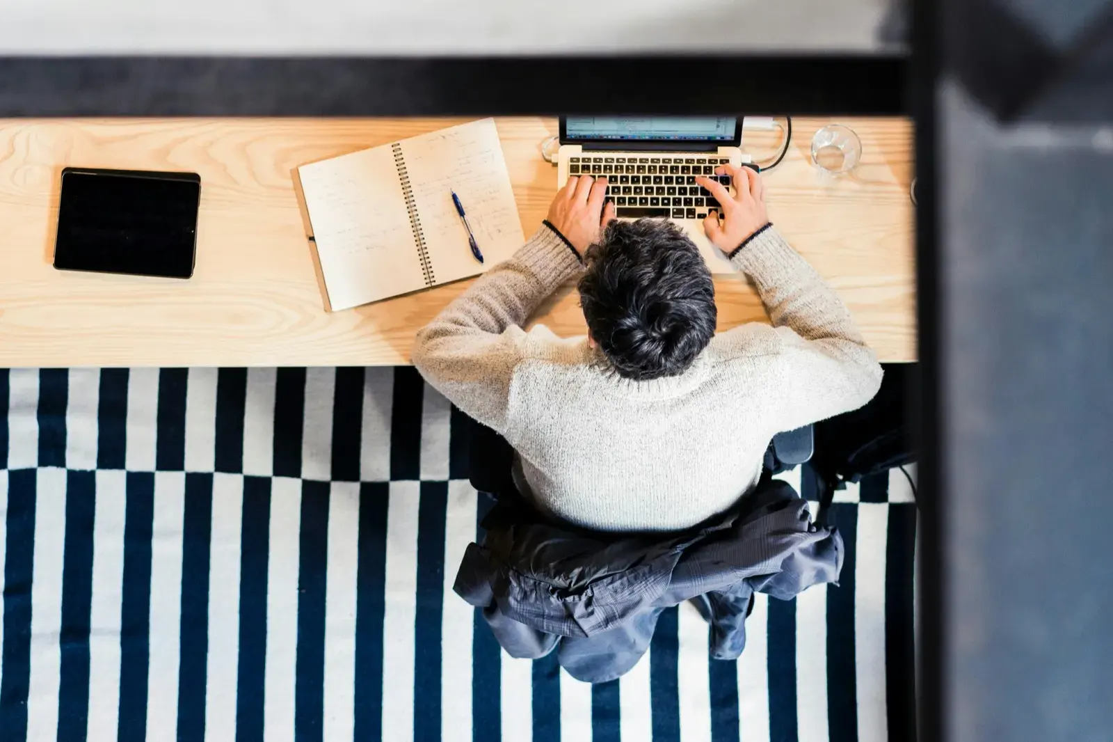 Person working at a desk with a laptop, notes, and tablet, viewed from above.